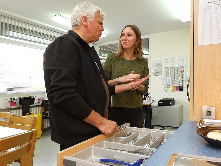 Ein Mann und eine Frau stehen in einer Küche an einer offenen Besteckschublade. Die Frau hält die linke Hand des Mannes und gibt ihm Lormen Zeichen mit ihrer Hand